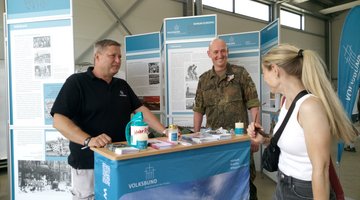 Kleiner Infostand mit zwei Männern, die mit einer Besucherin sprechen.